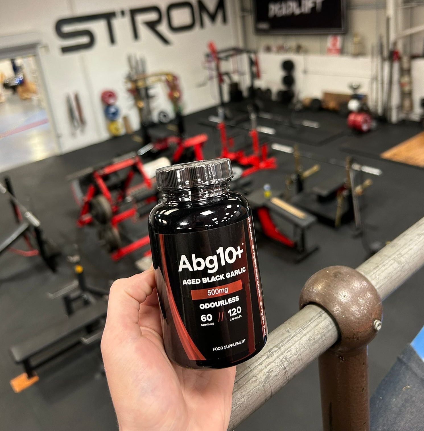 Hand holding a supplement bottle labeled 'Abg10+' in a gym setting with 'STROM' branding.